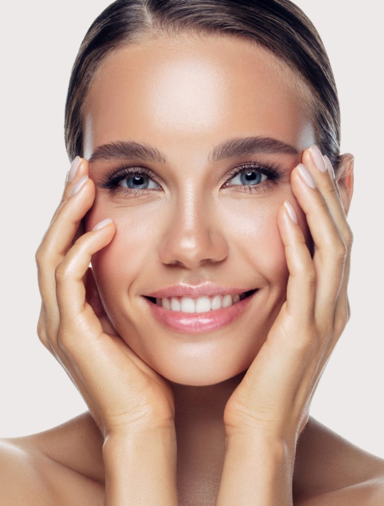 Smiling woman with clear skin gently touching her face against a light background.